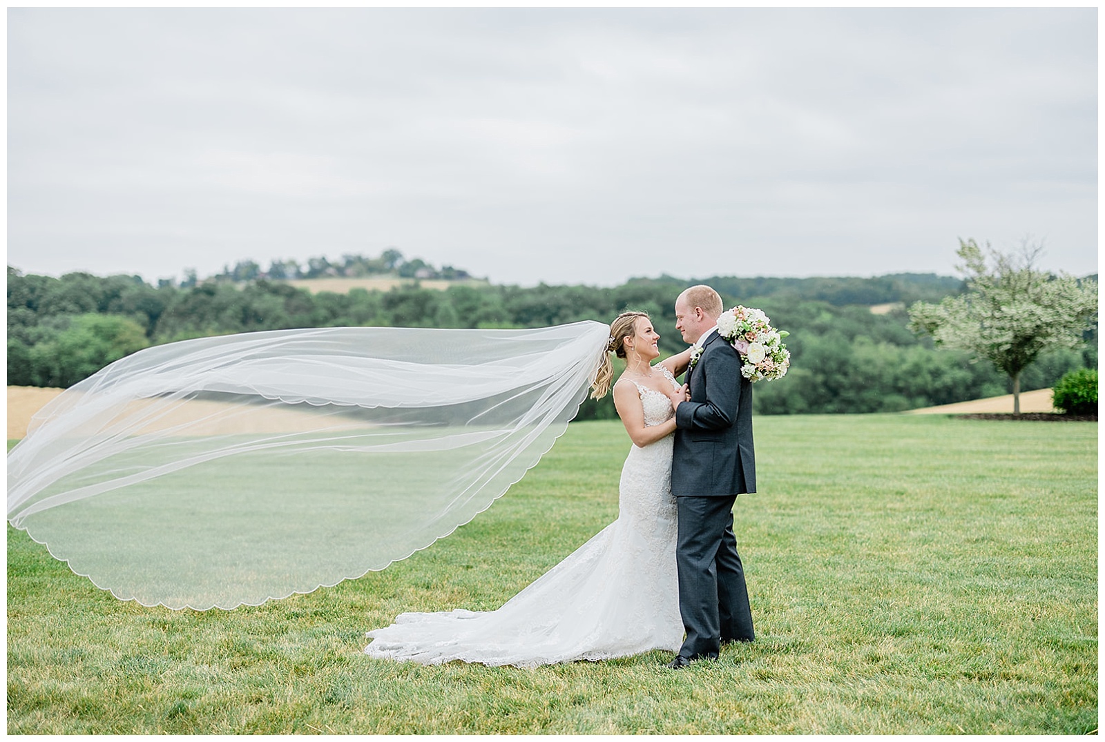 New bride and groom embrace in front of the pennsylvania mountain views at wyndridge farm wedding venue, with her long cathedral veil flowing in the wind
